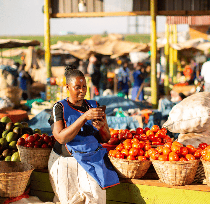 A woman wearing a blue apron sits at an outdoor market stall, holding a smartphone that may enable her business. She is surrounded by baskets filled with tomatoes and avocados, with blurred figures and stalls in the sunny background.