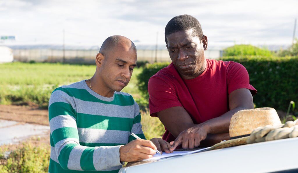 Two men stand outdoors by a car. One wears a green and grey striped jumper and writes on a clipboard, while the other, in a red shirt, observes seriously. A straw hat rests on the car, and a field with green crops is visible in the background.