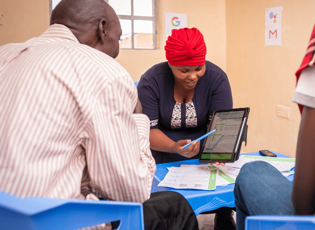A woman in a red headscarf smiles as she enables a man to learn, showing him a tablet and papers. They sit at a blue table with documents in a beige-walled room, where Google and Gmail logo posters hang in the background.