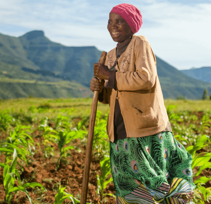 An elderly woman stands smiling in a lush green field, holding a wooden hoe - a testament to the harmony of tradition and agritech. She wears a pink headscarf, tan jacket, dark shirt, and a colorful patterned skirt, with mountains and blue sky behind her.