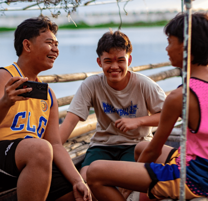 Fostering Human and Social Development at COP30: Three teenage boys sit together by the water, smiling and laughing. One wears a yellow basketball jersey and holds a phone showing COP30 news, while the others wear a gray T-shirt and a pink sports shirt with blue shorts.