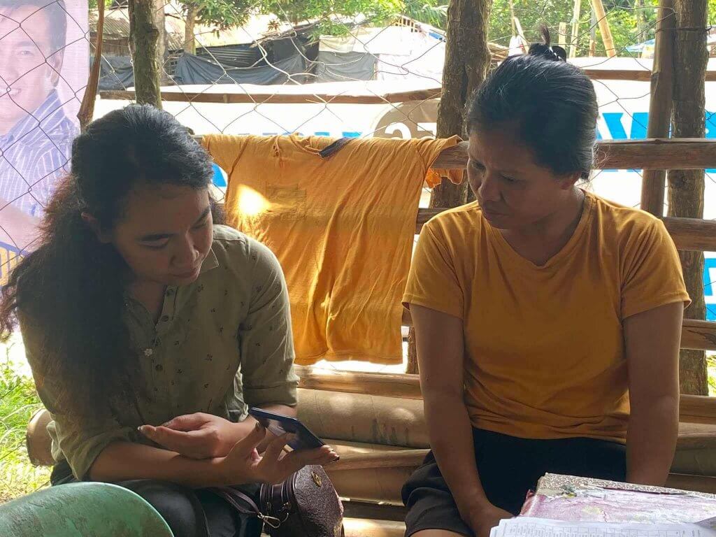 Two women sit on a wooden bench outdoors. The woman on the left, wearing a green shirt, shows something on her phone to the woman on the right, who wears a yellow shirt. A yellow shirt hangs on the fence behind them, and trees and other structures are visible.