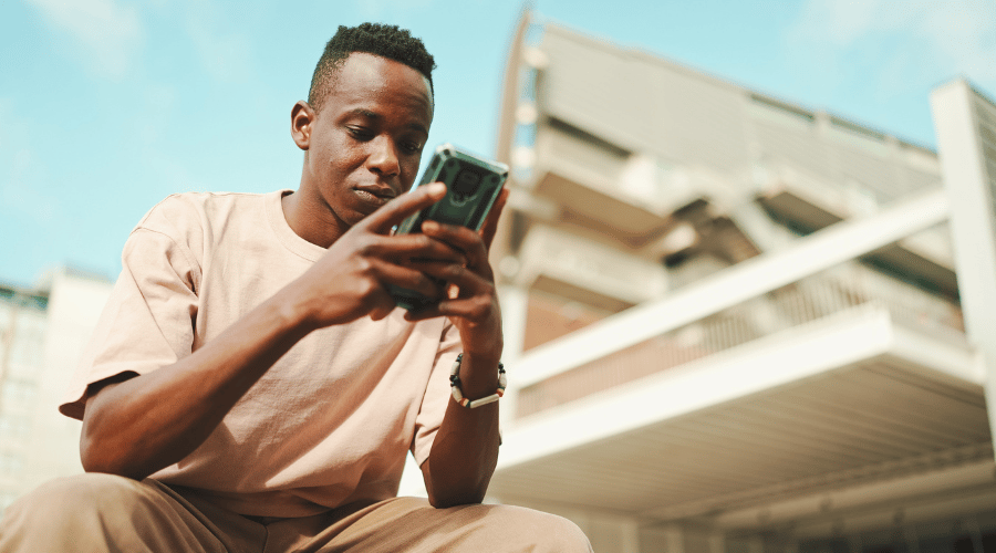 A young man wearing a light pink T-shirt sits outdoors, focused on his mobile phone. He is holding the phone with both hands. The background shows a modern, multi-storey building with balconies under a clear blue sky.