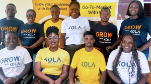 A group of ten smiling people, wearing "GROW with KOLA" and "KOLA MARKET" branded t-shirts in white, navy, and yellow, pose indoors in front of banners promoting KOLA Market and its go-to-market services. They sit and stand closely together.
