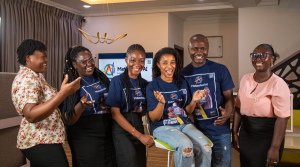 Six people stand indoors, smiling and laughing together. Four wear matching navy MetashoAI shirts, one wears a white floral blouse, and another wears a pale pink top with a black skirt. A TV shows the MetashoAI logo in the background. The setting is bright and modern.