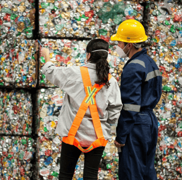 Two workers in safety gear and masks stand before bales of crushed cans and metal for recycling. As part of a ClimateTech initiative, one points ahead in a white coat and orange vest while the other, in blue, observes the process.