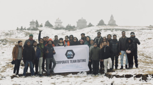 A group of people dressed in winter clothing pose outdoors on a snowy hillside, smiling and holding a banner that reads “CORPORATE TEAM OUTING.” In the background, there are stone structures and the sky appears overcast.