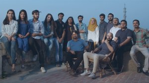 A group of fifteen young adults, both men and women, pose together outdoors at dusk. Some are sitting on chairs, others on a ledge, dressed in casual clothing. They smile calmly, with a tall tower and blue sky in the background.