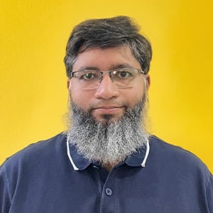A man with short dark hair and a full gray beard wearing glasses and a navy blue collared shirt with white trim stands in front of a solid bright yellow background, looking directly at the camera with a neutral expression.