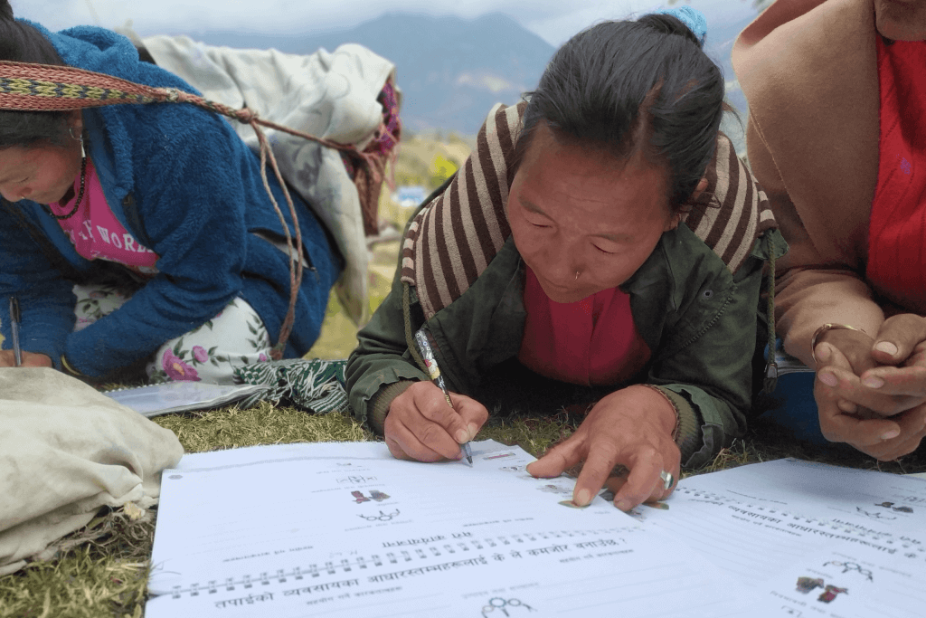 A woman lies on the grass in the Asia-Pacific, focused on writing in a workbook with Nepali script. She holds a pencil and wears a green jacket, whilst two others sit nearby. Mountains and a cloudy sky blur in the background.