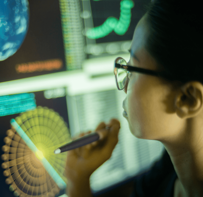A person with glasses closely examines data visualizations and charts on multiple computer screens, holding a pen near a circular graph—suggesting analysis or research in climate technology or science within a modern ClimateTech setting.