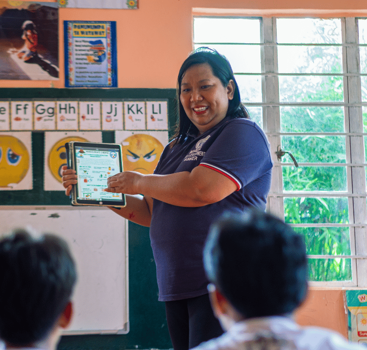 A smiling teacher in a navy blue shirt holds a tablet, showing its screen to students seated in a classroom. Alphabet posters, a green chalkboard, and utilities like sunlight streaming through the window brighten the learning space.