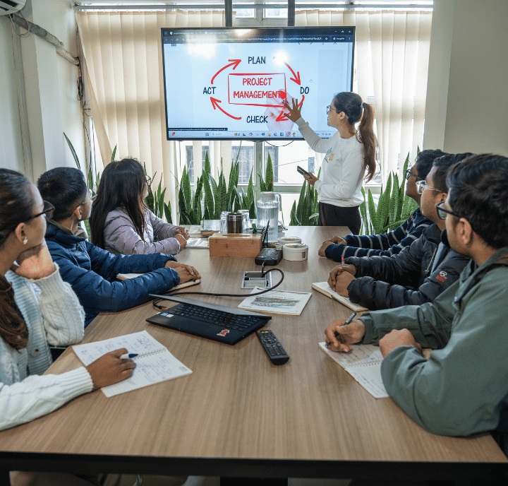 A woman stands by a screen showing a ClimateTech project management cycle diagram, explaining to six people seated at a conference table with papers, pens, and laptops in a bright office with tall plants and blinds.
