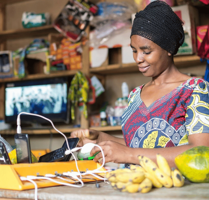 A woman in a patterned dress and headscarf smiles while charging several phones behind a wooden counter. Bananas and a watermelon are on the counter. Shelves with goods, a TV, and utilities hint at a lively small shop setting.