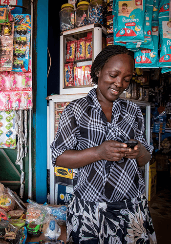 A woman in a plaid shirt stands smiling and looking at her phone inside a small shop. Shelves behind her display colorful toys, snacks, and Pampers diapers. The shop is packed with various goods in bright packaging.