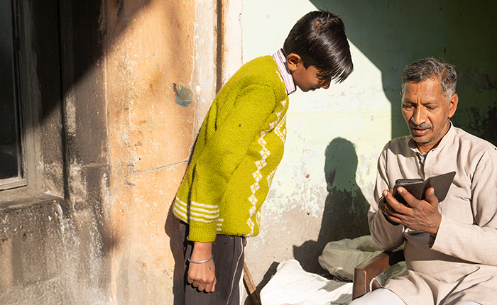 A man in light traditional clothing sits on a chair outdoors, holding a smartphone. A boy in a green sweater leans over to look at the screen. Sunlight casts strong shadows on the old, weathered wall behind them.