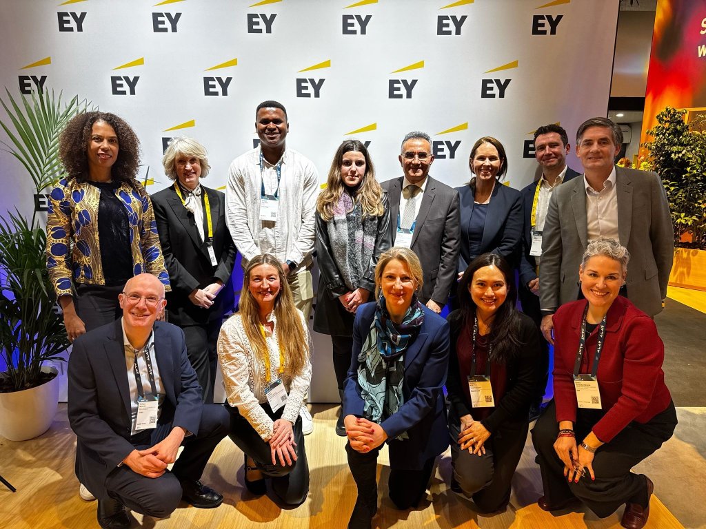 Thirteen professionally dressed people pose and smile in two rows in front of an EY (Ernst & Young) branded backdrop. Some are kneeling in the front; others stand behind them. There are green plants on both sides of the group.