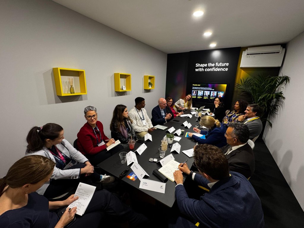 A group of professionals, diverse in gender and ethnicity, sit around a rectangular table in a modern meeting room. They are engaged in discussion, taking notes, and listening. The wall has a screen displaying "Shape the future with confidence.