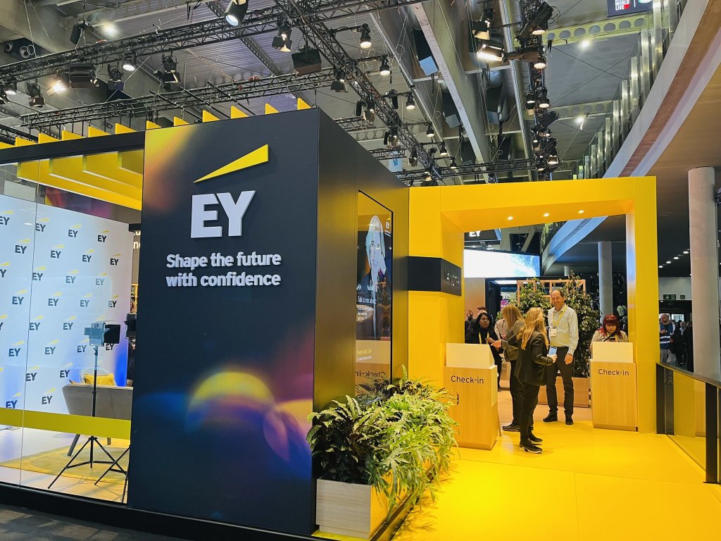 A modern EY exhibition stand with yellow and black design, featuring the EY logo and the slogan "Shape the future with confidence." Several people stand near a check-in counter, and the area is well-lit with hanging lights and plants for decoration.