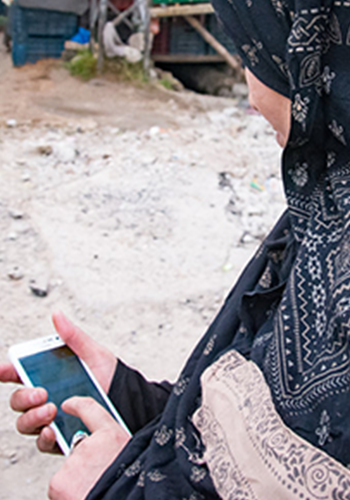 A person wearing a patterned black and beige hijab holds a white smartphone with both hands. The screen reflects their fingers. The background is outdoors, with a dirt ground and some blurred objects and structures. The person's face is not visible.