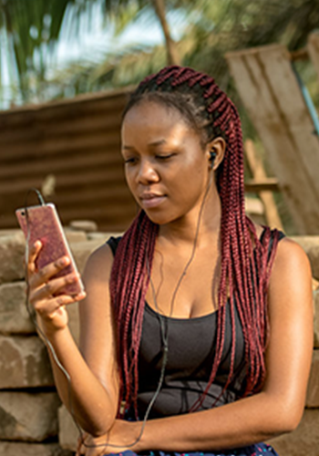 A young woman with long red braids sits outdoors, wearing earphones and looking at her smartphone. She is dressed in a black sleeveless top, with a background of stacked bricks, wooden planks, and green foliage.