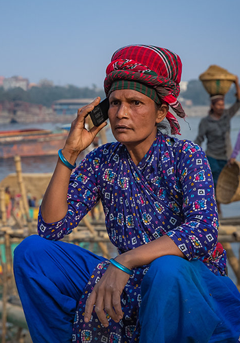 A person wearing a patterned blue outfit and a red-checkered headscarf sits outdoors, holding a cellphone to their ear. The background shows blurred people working, bamboo structures, river, and boats, suggesting a busy riverside or market scene.