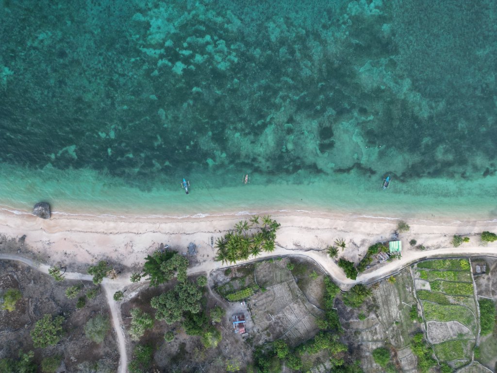 Aerial view of a beach with turquoise water, a sandy shoreline, and small boats near the shore. Tropical trees surround the area, while dirt paths and agricultural plots hint at the Central Insights Unit’s efforts beyond the sand.