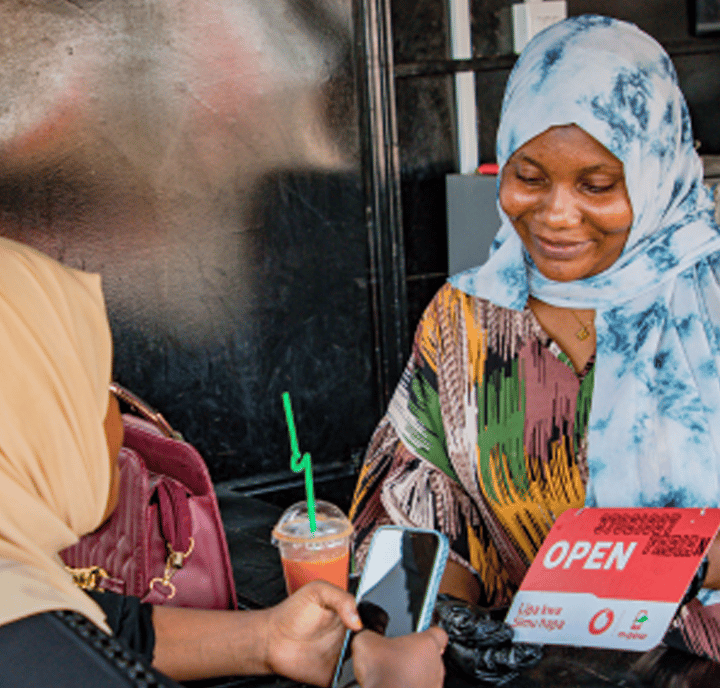 Two women sit together at a table. One, in a blue patterned hijab, smiles and holds an “OPEN” sign—a symbol of welcoming public policy. The other, in a beige hijab, checks her smartphone. A pink drink with a green straw rests nearby.