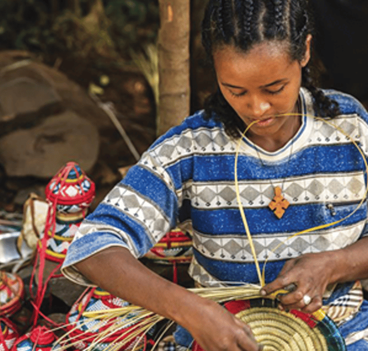 A woman with braided hair, wearing a blue and white patterned shirt and a wooden cross necklace, weaves a colorful basket from straw. Her artistry highlights the importance of PRA in supporting traditional crafts and empowering artisans.