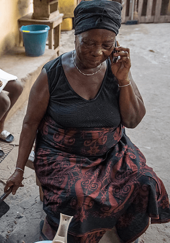 An elderly woman wearing a black headscarf, sleeveless black top, and patterned wrap skirt sits on a stool outdoors, smiling while talking on a mobile phone. A blue bucket and a wooden bench are visible in the background.