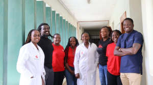 Eight people stand in a hallway, smiling at the camera. Two wear white lab coats, four wear matching red shirts, one wears a navy shirt with a logo, and one wears black. The corridor has green pillars and off-white walls.