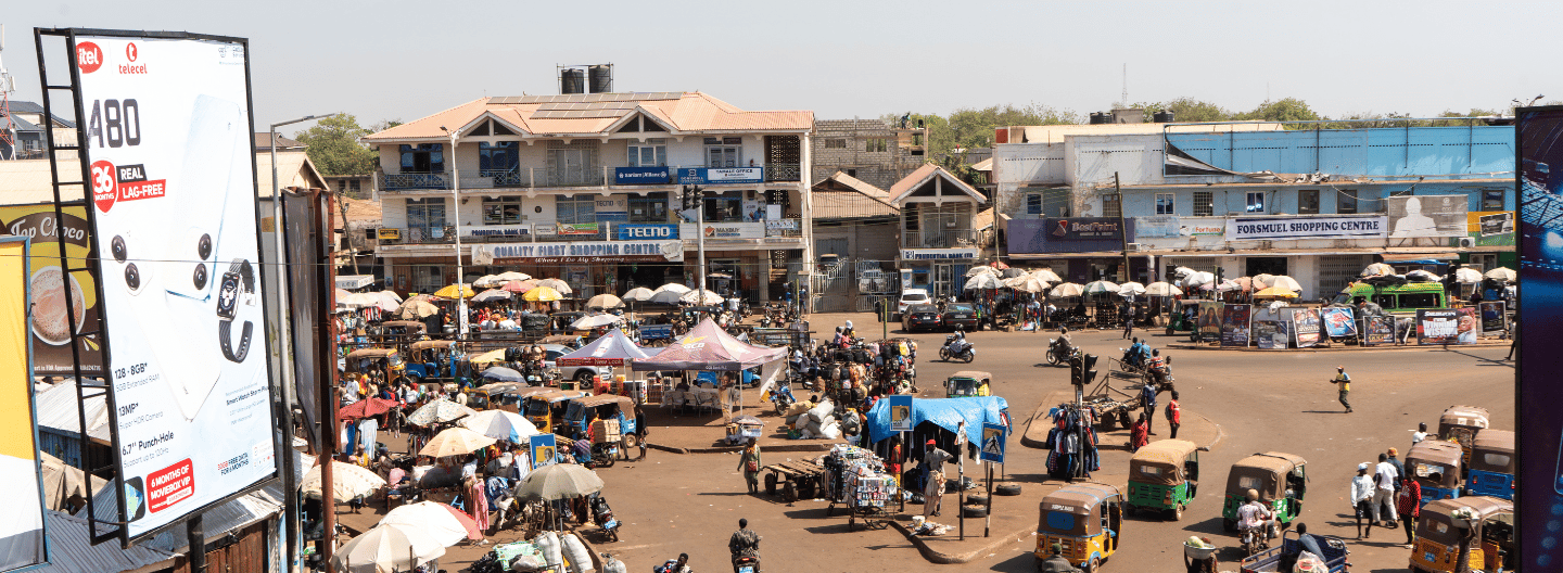 A bustling outdoor market scene in an urban area, with many people, motorcycles, auto rickshaws, and street vendors under umbrellas. Low-rise buildings feature visible utilities infrastructure, while large billboards frame both sides of the image.