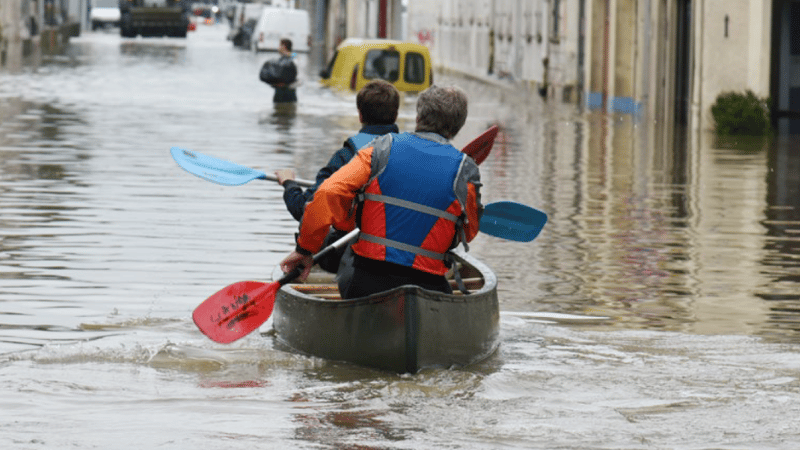 Two people in life jackets paddle a canoe down a flooded city street, where mobile-enabled early warning systems could have provided alerts. Water submerges the road as others and vehicles struggle in the background, with buildings lining both sides.