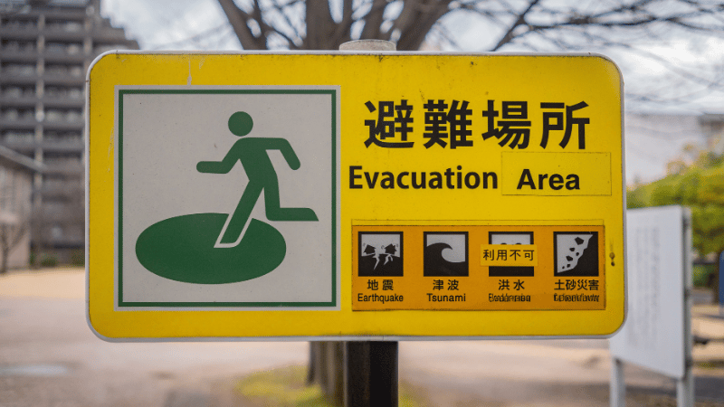 A yellow sign reads “Evacuation Area” in English and Japanese, with a running figure icon. Below are icons for earthquake, tsunami, flood, and landslide hazards—supported by mobile-enabled early warning systems—and a symbol indicating restrictions.