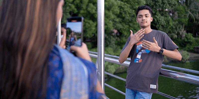 A young man in a graphic t-shirt poses with hand signs beside a metal railing on a bridge over green water. A person with long brown hair and a blue shirt is in the foreground, taking his picture with a mobile phone. Trees fill the background.