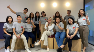 A group of thirteen people, smiling and posing together indoors. Most wear matching gray shirts with a small red heart on the chest. Some are seated on wooden blocks, while others stand behind. The background is plain with three round lights on the wall.