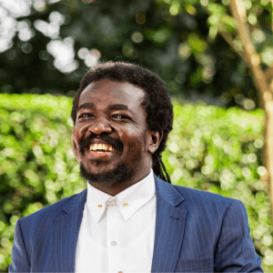 A man with medium-length curly hair and a beard smiles broadly while wearing a blue pinstripe suit jacket and a white dress shirt. He stands outdoors in front of green foliage with sunlight filtering through the trees.