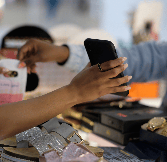 A close-up of two hands during a retail transaction. One hand holds a phone to scan a QR code on a small shopping bag, highlighting digital payments in retail—a subtle nod to innovation in areas like Climate Finance amid sandals and accessories softly displayed.
