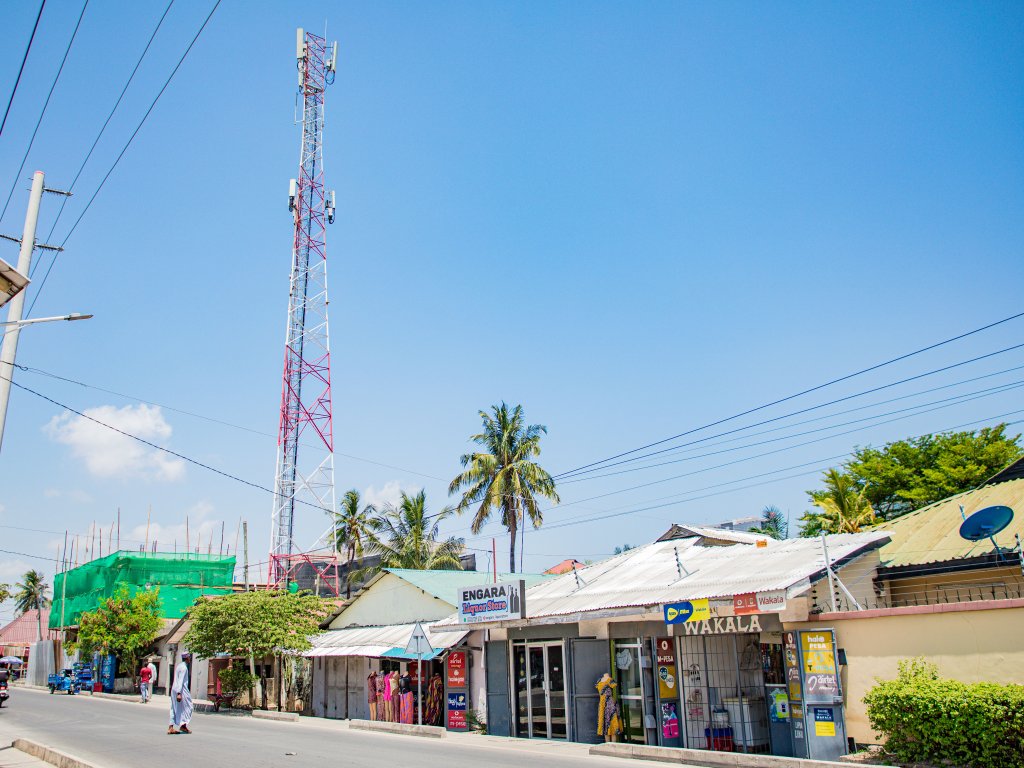 A street scene under a bright blue sky shows small shops with metal roofs, a tall red and white mobile mast, palm trees, power lines, and a few pedestrians walking on the pavement. Signs on shopfronts read "ENGARA" and "WAKALA.