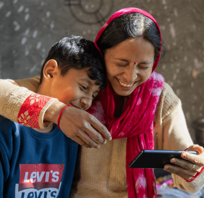A smiling woman in a red scarf and traditional attire sits closely with a laughing boy in a blue Levi’s sweatshirt. Both look at a smartphone, sharing a joyful moment as they read about climate finance in the softly lit room.