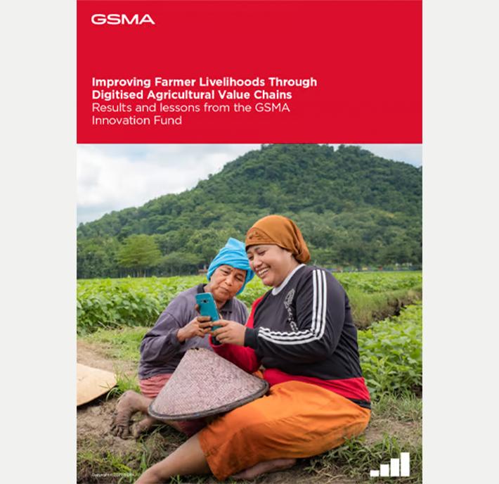 Two women sit outdoors in a lush, green rural area with hills in the background. One elderly woman wears a blue shirt and headscarf, while the younger woman in a red skirt and orange headscarf smiles as she shows something on a mobile phone to the elder.