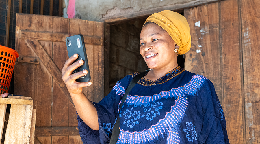 A smiling woman in a blue patterned dress and yellow headscarf holds up a mobile, appearing to take a selfie or video call. She stands in front of a rustic wooden door, with a red plastic basket and wooden shelves visible nearby.