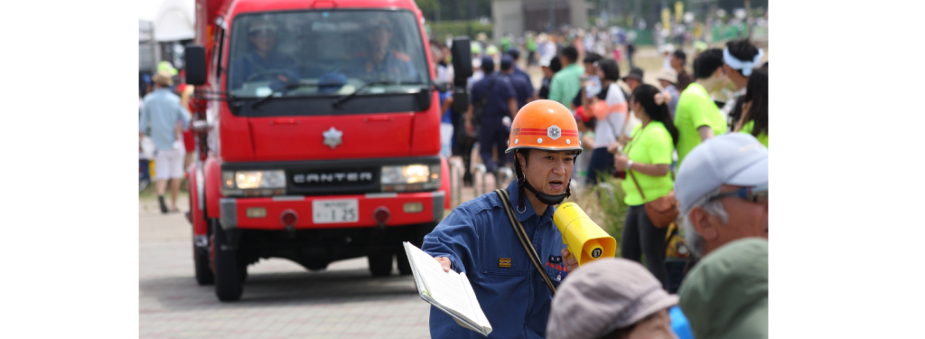 Firefighter with a helmet gives early warning instructions using a megaphone; a fire engine and crowd are in the background.