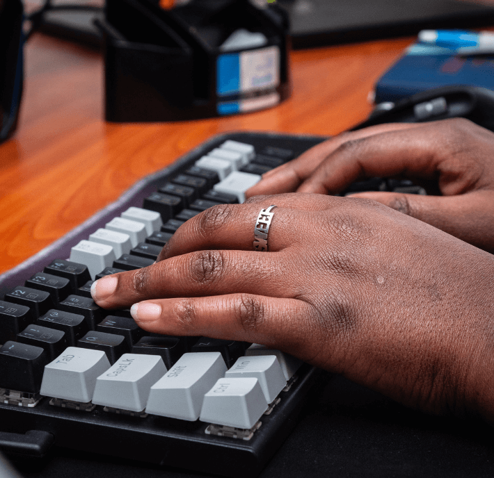A close-up of dark-skinned hands typing on a black-and-white computer keyboard, possibly drafting a report on Climate Finance. One hand wears a silver ring with engraved text. The background features a wooden desk and blue notebook, slightly out of focus.
