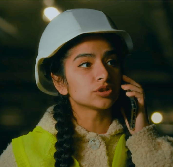 A young woman wearing a white safety helmet and yellow high-visibility vest talks on her mobile phone, appearing focused. Indoors with blurred lights in the background, she embodies digital humanitarian assistance in action.