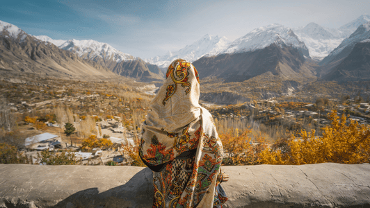 A person in a patterned shawl looks at snow-capped mountains and an autumn valley landscape.