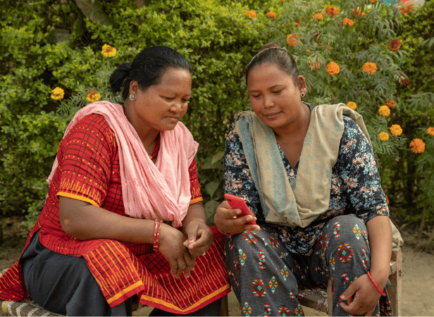 Two women sit outdoors, looking at a mobile phone together, surrounded by green plants and orange flowers—highlighting the real-world impact featured in the Mobile for Humanitarian Innovation Annual Report 2025.