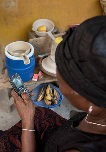 A woman wearing a headscarf uses a mobile phone near a blue water container and a bowl of bananas.