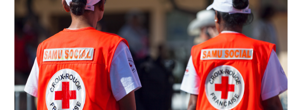 Two people wearing orange SAMU SOCIAL vests with French Red Cross logos, seen from behind, coordinate Early Warnings to support emergency response efforts.