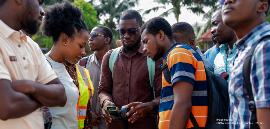 A group of people outdoors examining a handheld device together, with trees in the background—a scene reflecting collaboration highlighted in the Mobile for Humanitarian Innovation Annual Report 2025.
